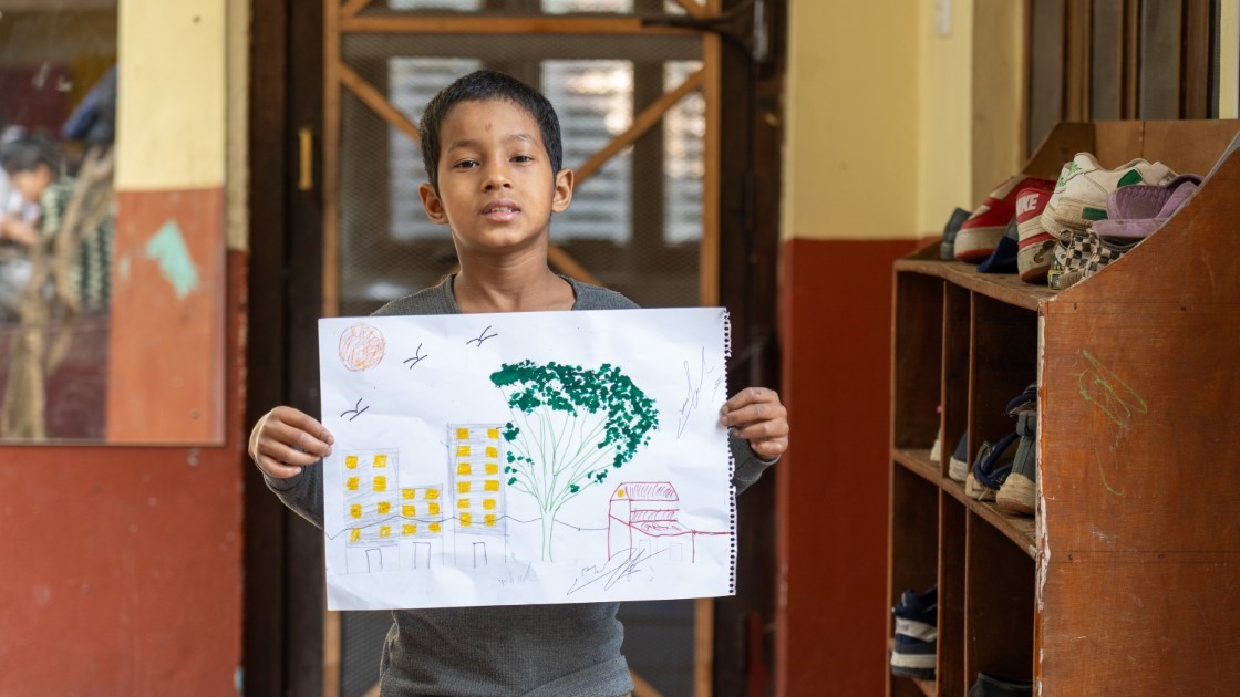 A young boy stands indoors holding up a drawing. The artwork depicts a sun, birds, buildings, a tree, and a bench. Behind him is a wooden shoe rack filled with various shoes. The background features a glass door and a reflection.