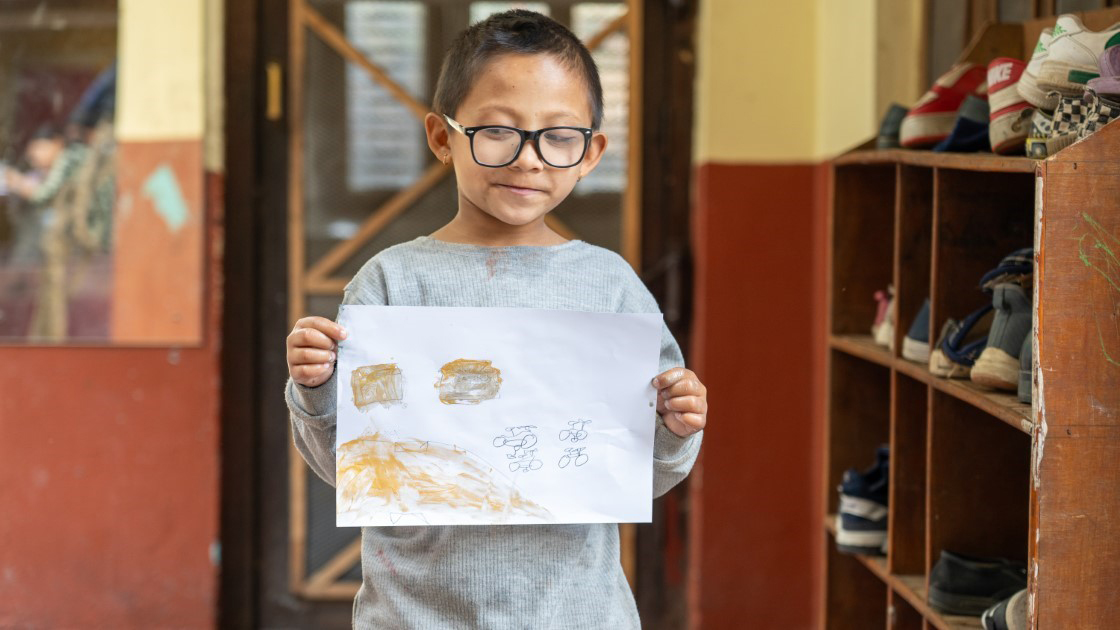 A child with glasses holds up a sheet of paper displaying a drawing of bicycles and houses. The child is standing indoors near a shoe rack, and there are shoes on the shelves.