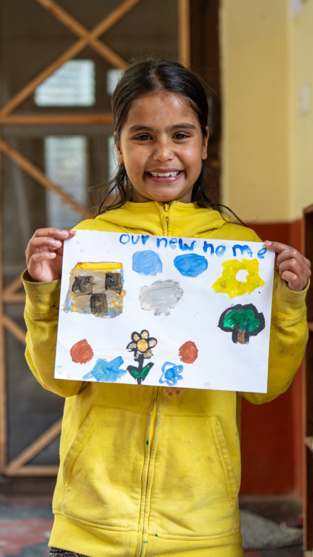 A smiling child in a yellow hoodie holds a colorful drawing with a house, flowers, trees, and sky. The artwork is titled "Our New Home." They stand indoors, in front of a door with a visible doormat.
