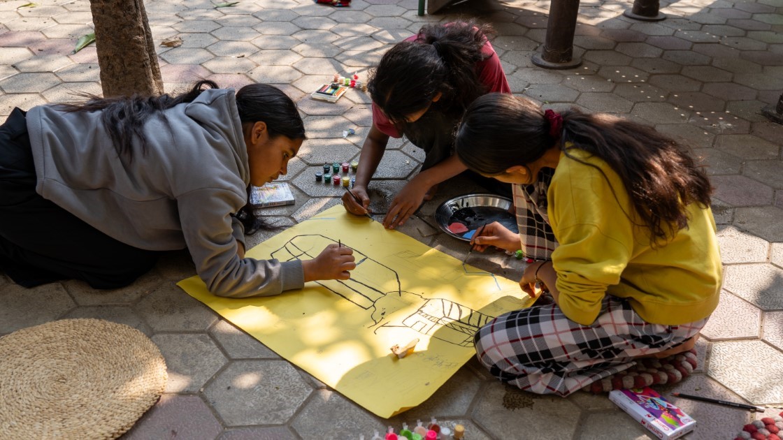 Three children are drawing on a large yellow paper laid on a paved surface. They are surrounded by crayons and art supplies. They appear to be focused on their artwork. Sunlight casts shadows across the scene.