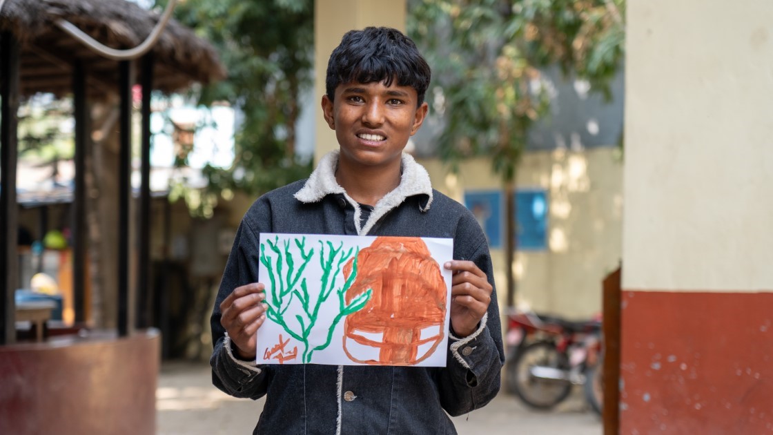 A person in a denim jacket stands outdoors, holding up a drawing of a tree and a brown shape. The background includes trees, a bike, and a building.