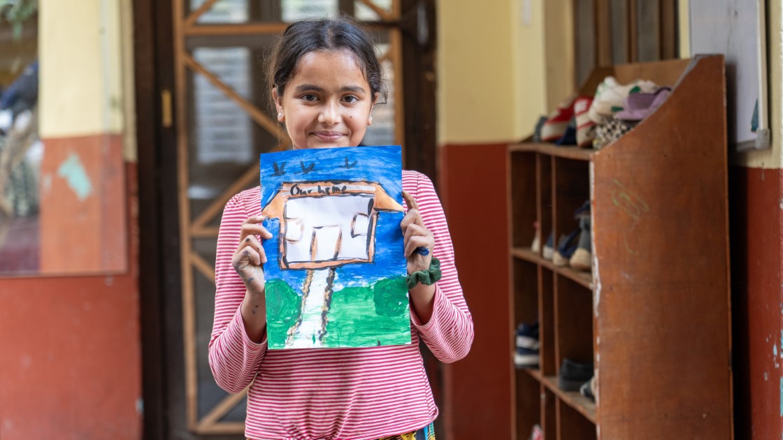 A girl in a striped shirt holds up a colorful drawing of a house, smiling proudly. She stands in a hallway with wooden shelves holding shoes in the background.