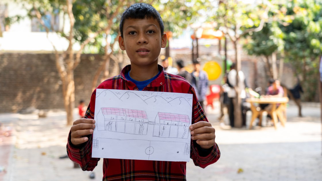 A young boy in a red plaid shirt holds up a drawing of a house with mountains in the background. He is outdoors in a sunlit area with blurred trees and people behind him.
