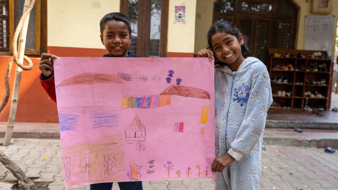 Two smiling children stand outside holding a large, colorful drawing. The background features a building with windows and a shoe rack. The pavement is made of gray bricks.