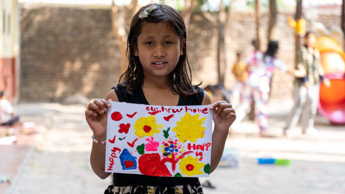 A young girl stands outdoors, holding a colorful drawing with flowers and the words "Khushiyon Aur Hum Home Happy." She wears a black top and has a hair clip. Blurred background shows people and play equipment.