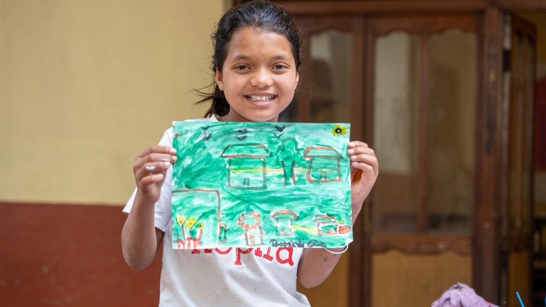 A young girl smiles and holds up a colorful drawing depicting houses and trees. She is wearing a white shirt and standing indoors against a wooden backdrop.
