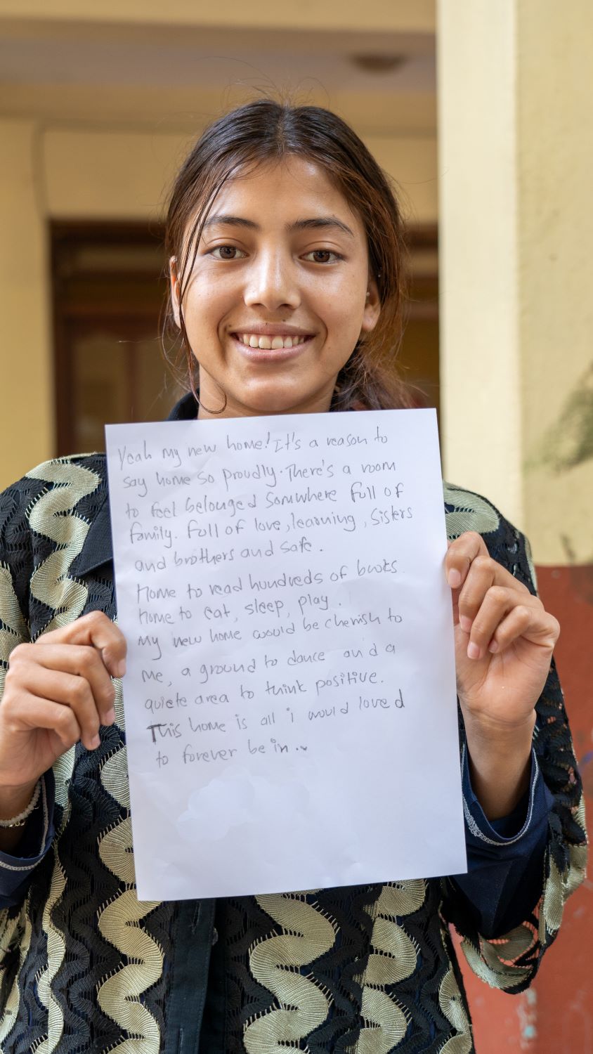 Smiling person holding a handwritten note. The note mentions feelings of comfort, belonging, and love for a new home, describing it as a place for family, rest, and positivity. The background is a simple indoor setting.