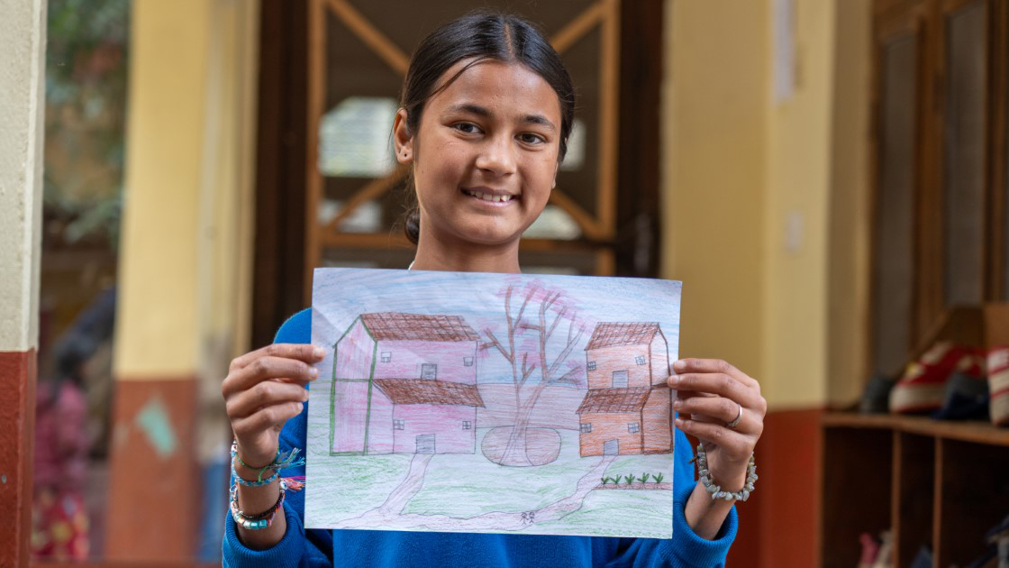 A young girl smiles while holding up her drawing of two houses with red roofs, a tree, and pathways. She stands indoors, and shoes are visible in the background.