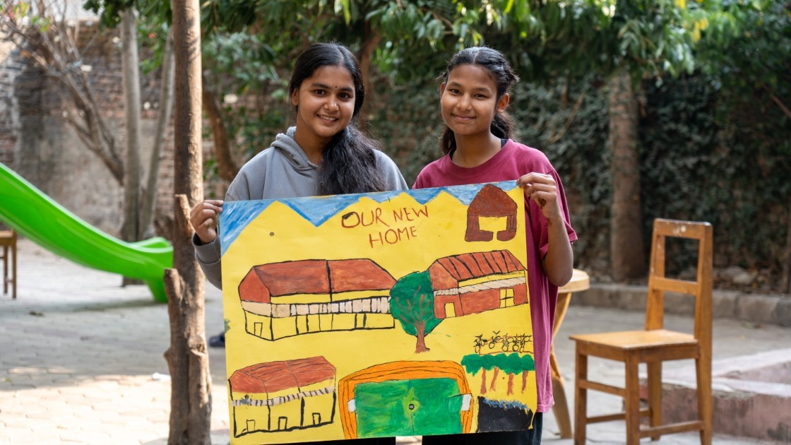 Two girls stand outdoors holding a colorful drawing titled "Our New Home," depicting houses and trees. They are smiling, with a playground slide and chairs visible in the background, surrounded by greenery.