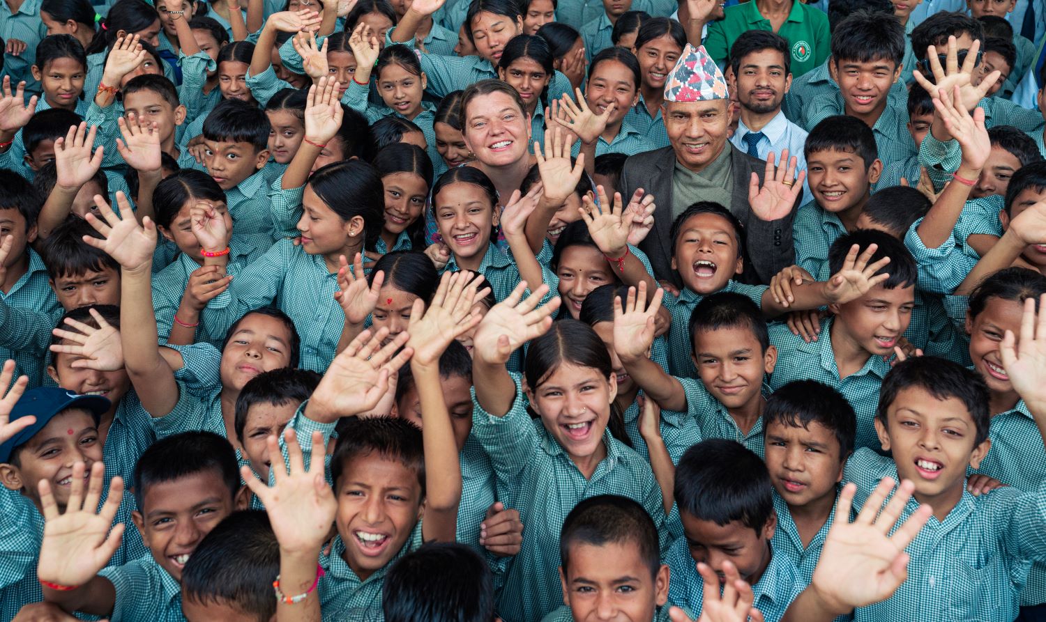 A large group of smiling children in school uniforms raise their hands enthusiastically. Among them are a few adults who are also smiling. They all appear joyful and are standing closely together.