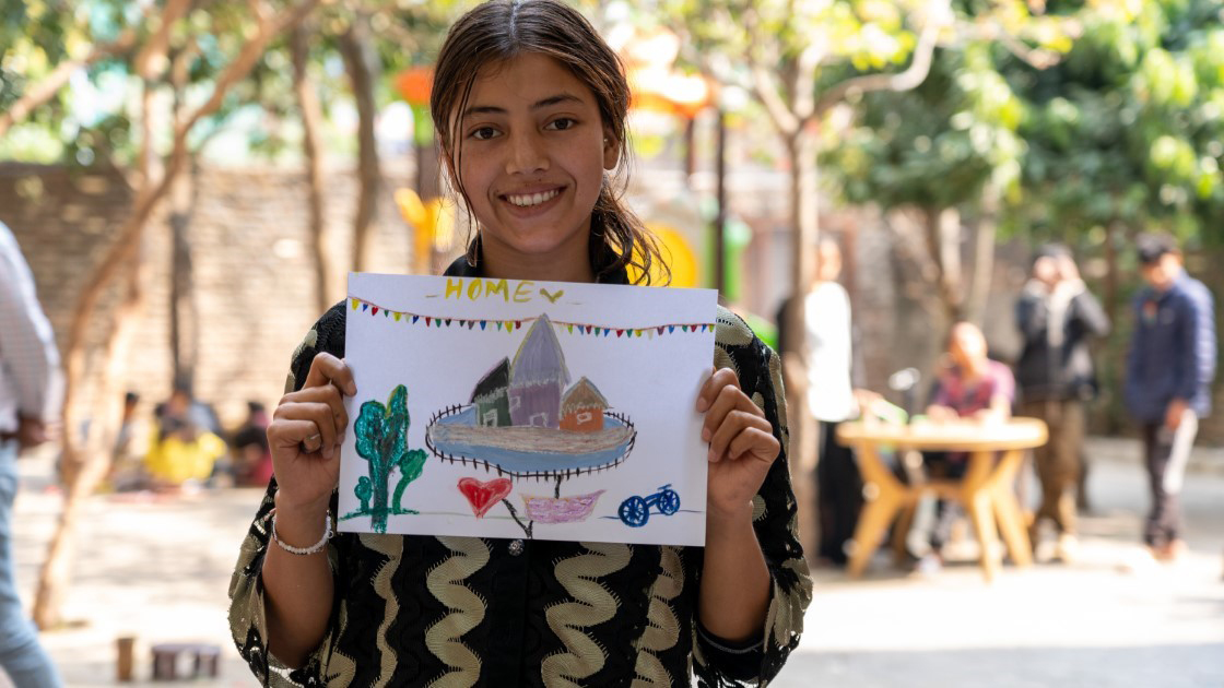 A young person is smiling and holding a colorful drawing labeled "Home," depicting a house, trees, and a heart. They stand outdoors with blurred people and trees in the background.