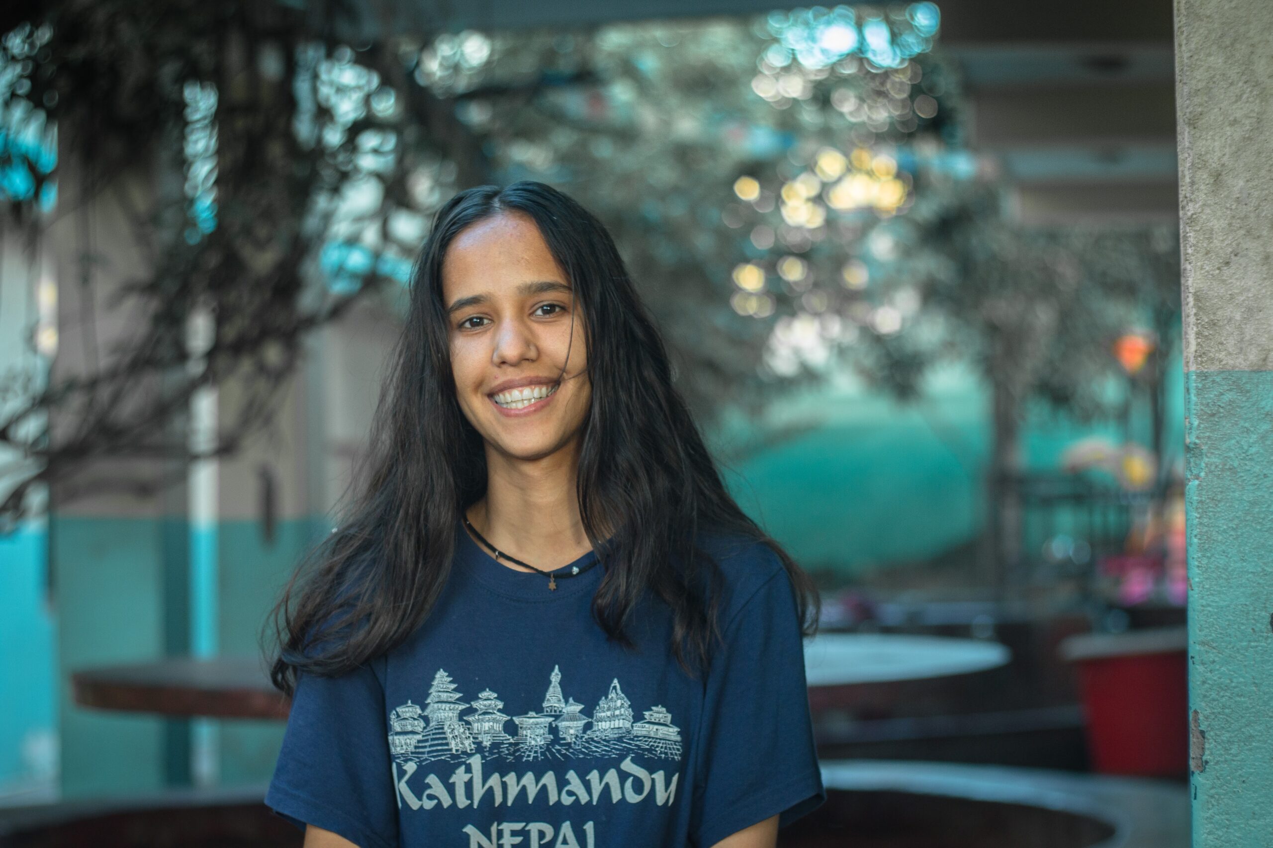 A young woman with long dark hair smiles while standing outdoors, wearing a navy blue "Kathmandu Nepal" t-shirt. The background is blurred with trees and a building visible.
