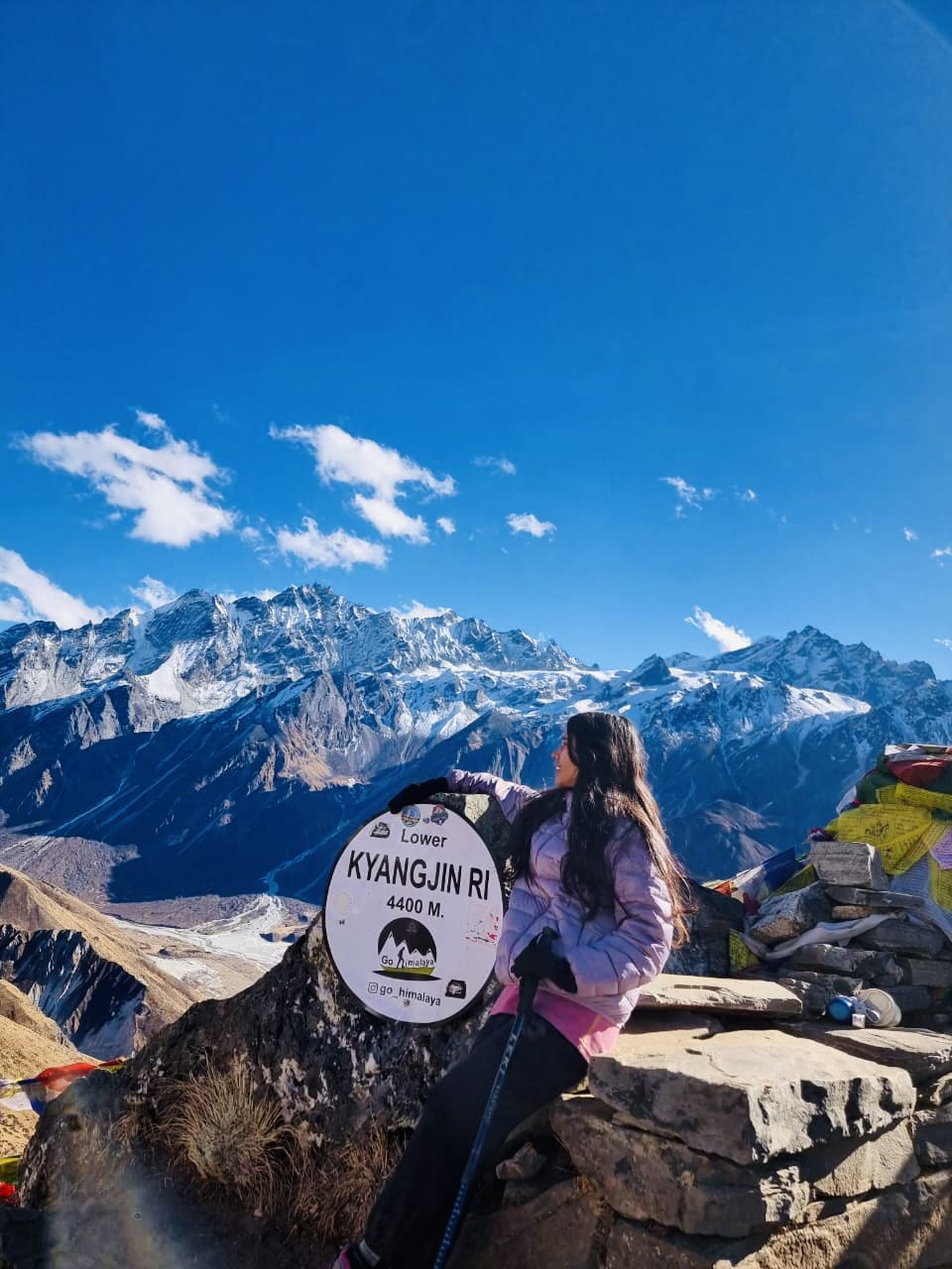 A woman in a light purple jacket sits on a rock next to a "Lower Kyangjin Ri 4400 M" sign, with snow-capped mountains and a bright blue sky in the background.