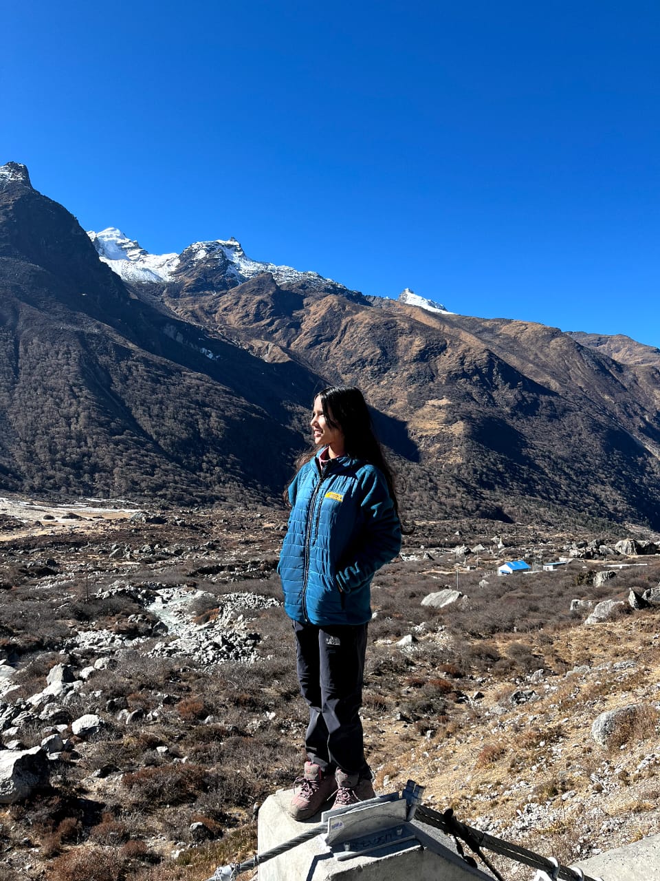 A woman in a blue jacket stands on a rock, looking into the distance with snow-capped mountains, rugged hills, and a clear blue sky in the background.