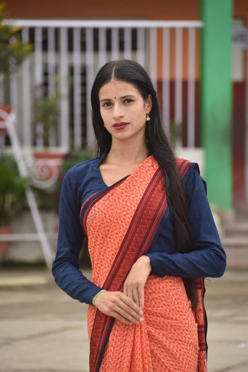 A woman wearing a patterned orange saree with a navy blue blouse stands outdoors, looking at the camera with a calm expression. She has long black hair and traditional jewelry, and the background is blurred.