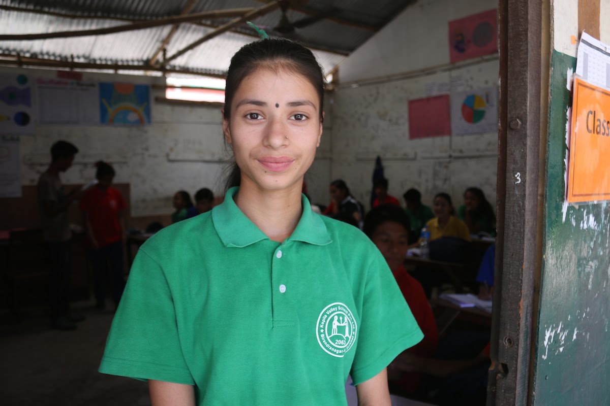 A young woman wearing a green school uniform stands in a classroom doorway, smiling at the camera. Other students are seated inside, with colorful charts on the walls. A "Class" sign is visible on the door.