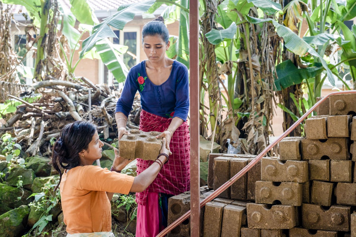 Two women outdoors, with one handing a large brick to the other. They are surrounded by stacked bricks and lush green plants, working together near a pile of branches and a building in the background.