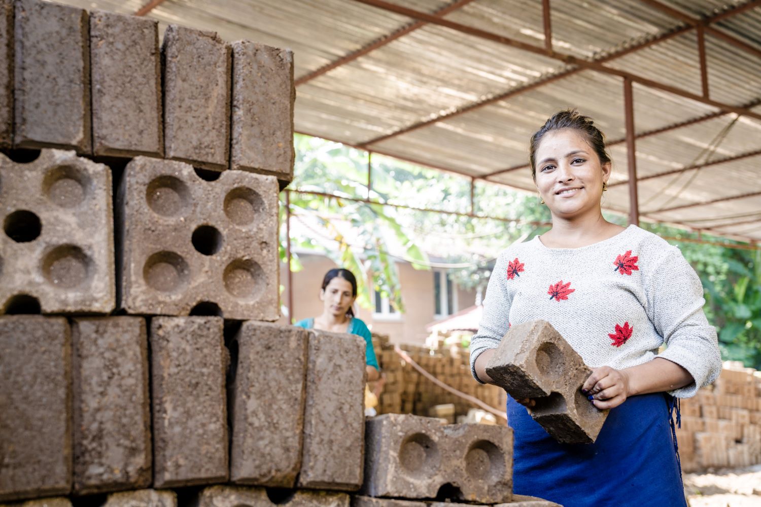 A woman smiles while holding a brick in a covered outdoor area with stacks of bricks. Another woman stands in the background, partially visible. The setting appears to be a brick-making or construction site.