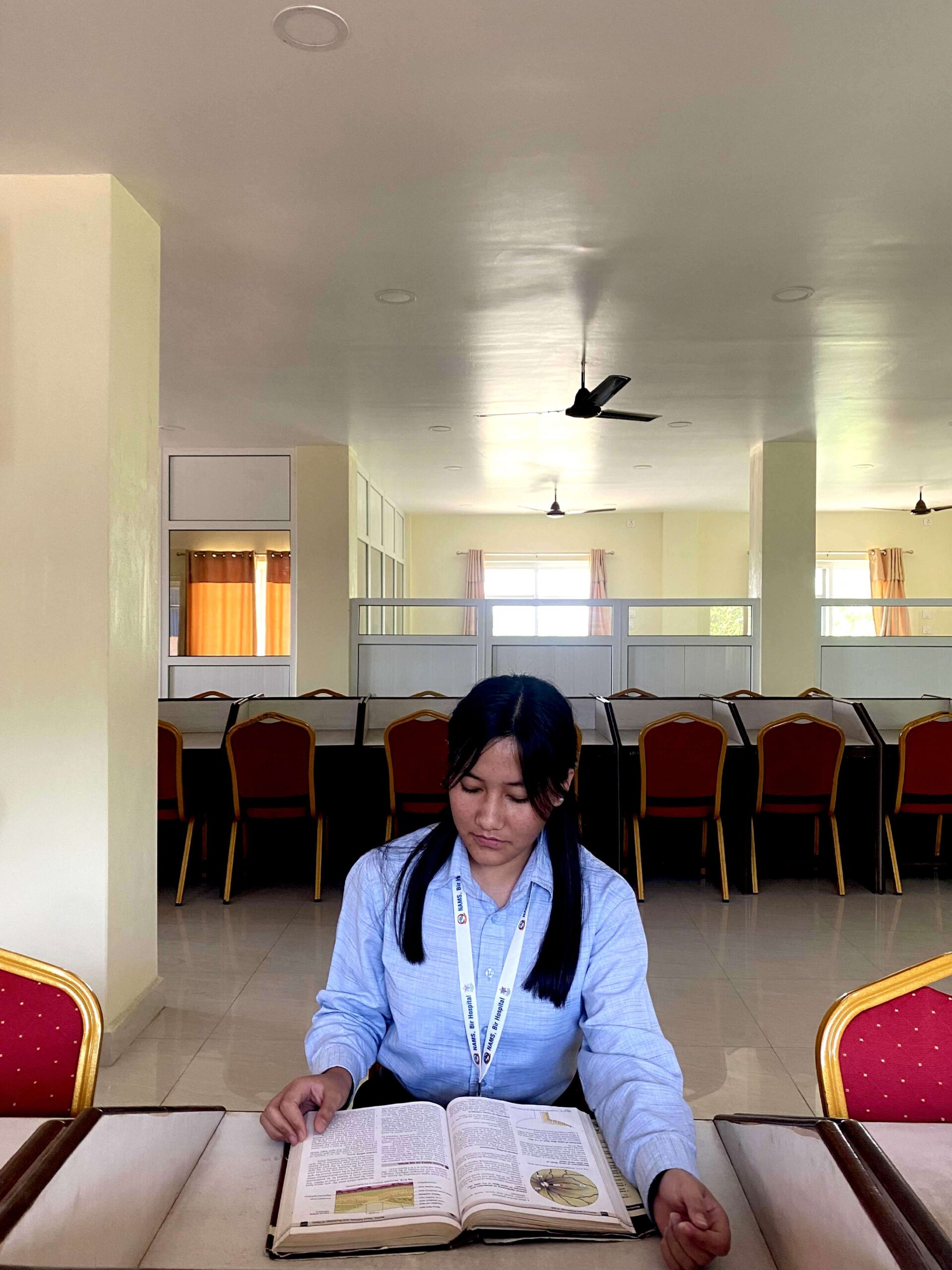 A young woman with long dark hair and a lanyard sits alone at a table in a bright, spacious room, reading an open book. Red chairs are arranged in the background.