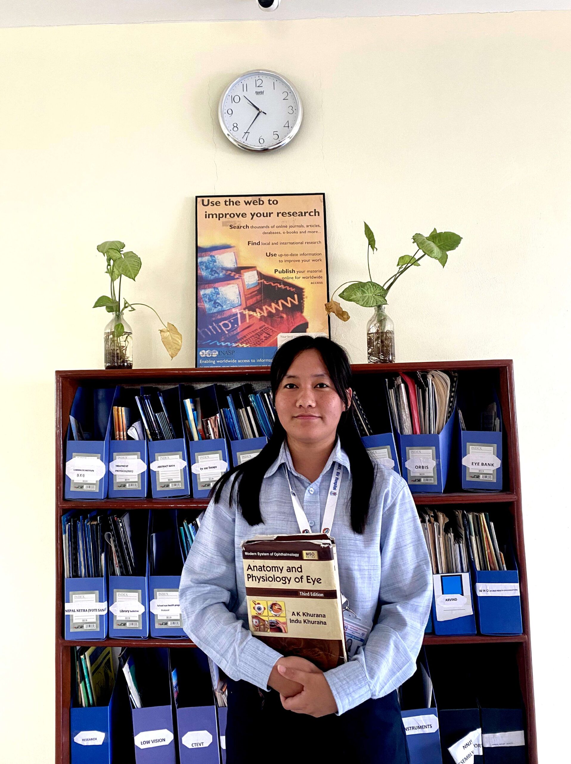 A woman stands in front of a bookshelf filled with labeled blue and yellow folders, holding two books. There is a wall clock and a research poster above her, with two plants in glass jars on the shelf.