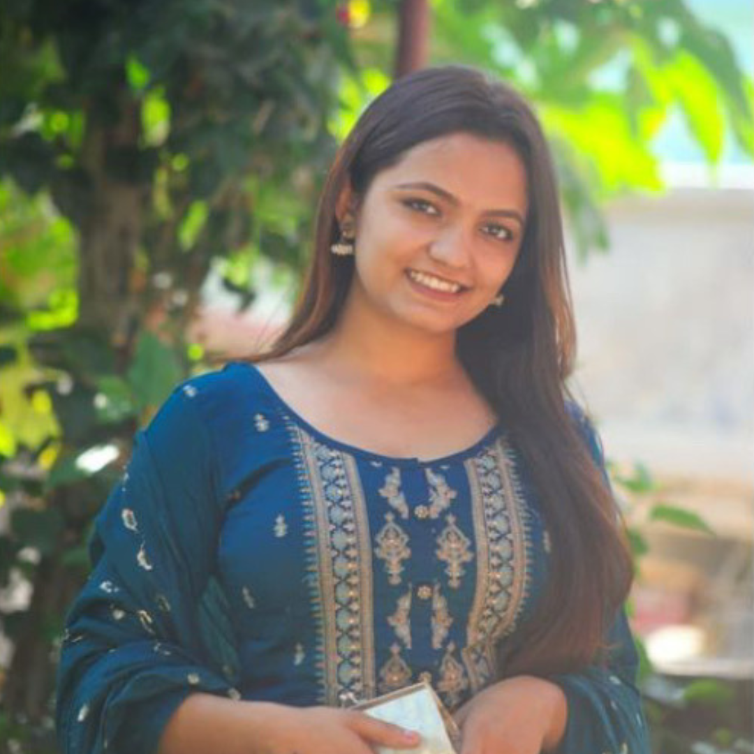A young woman with long brown hair smiles while posing outdoors in a blue traditional outfit with gold embroidery, holding a small purse; green foliage is visible in the background.
