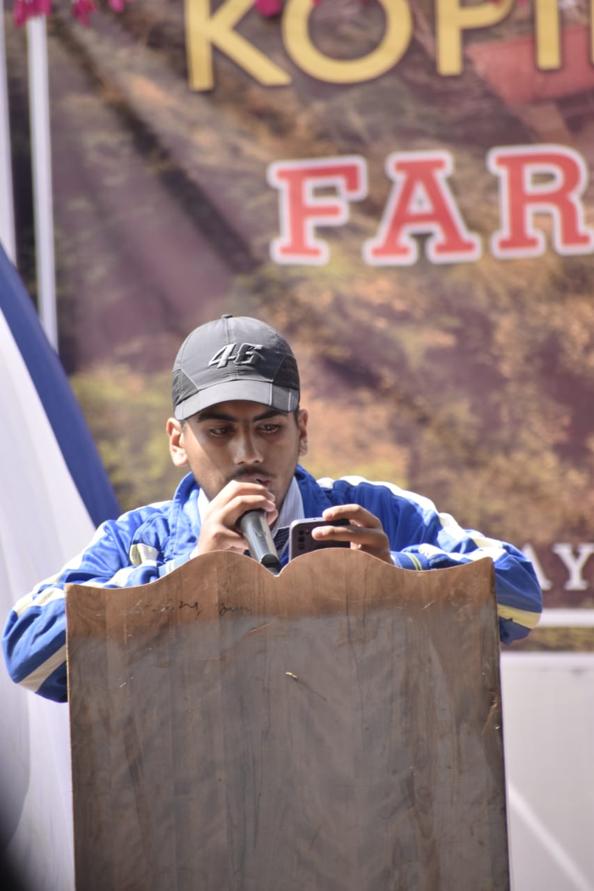 A young man wearing a black cap and blue jacket speaks into a microphone while looking at his phone behind a wooden podium at an outdoor event. A banner with bold text is visible in the background.