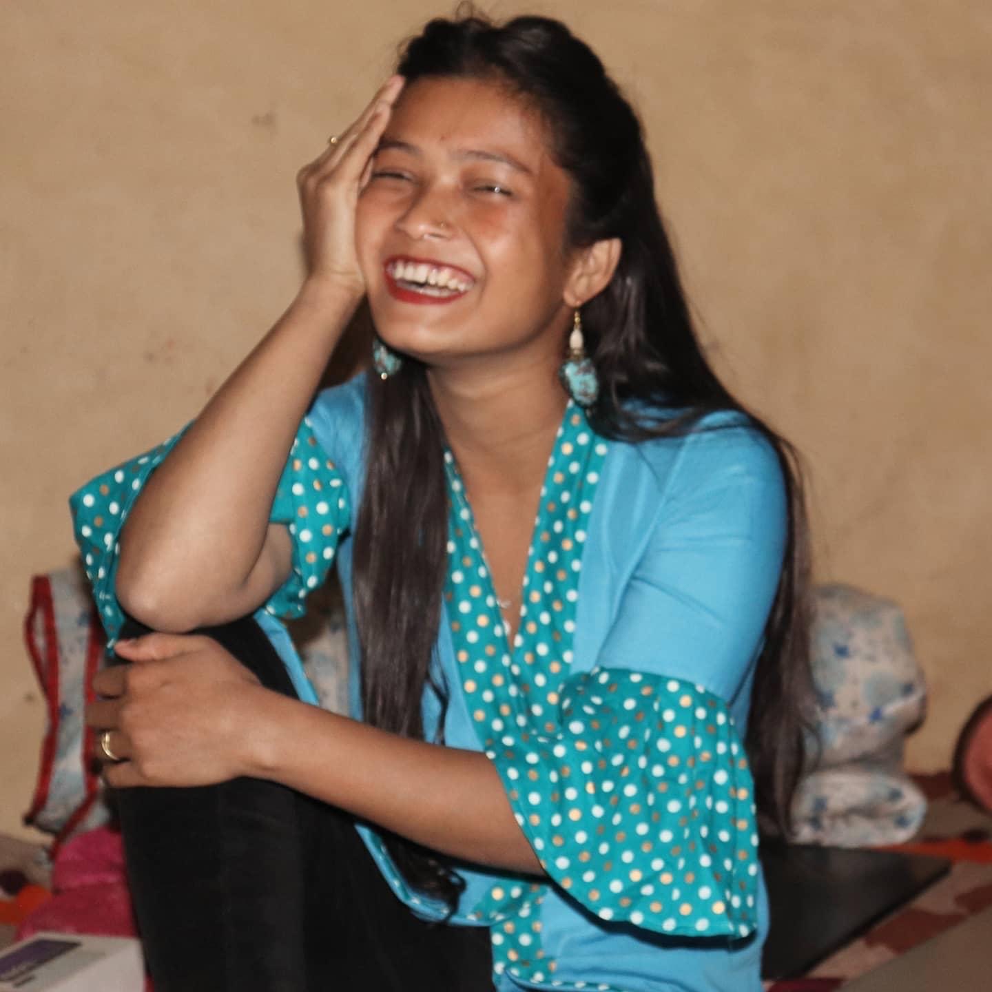 A young woman with long dark hair, wearing a blue top with polka dots, sits cross-legged and laughs joyfully with her hand on her face, against a plain beige background.