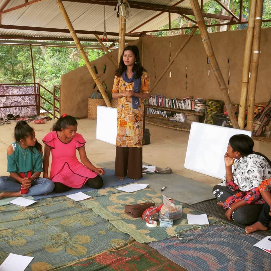 Four women sit on rugs with papers around them, while another woman stands and speaks. They are in an open, bamboo-structured room with books, whiteboards, and natural light, suggesting a workshop or group activity.