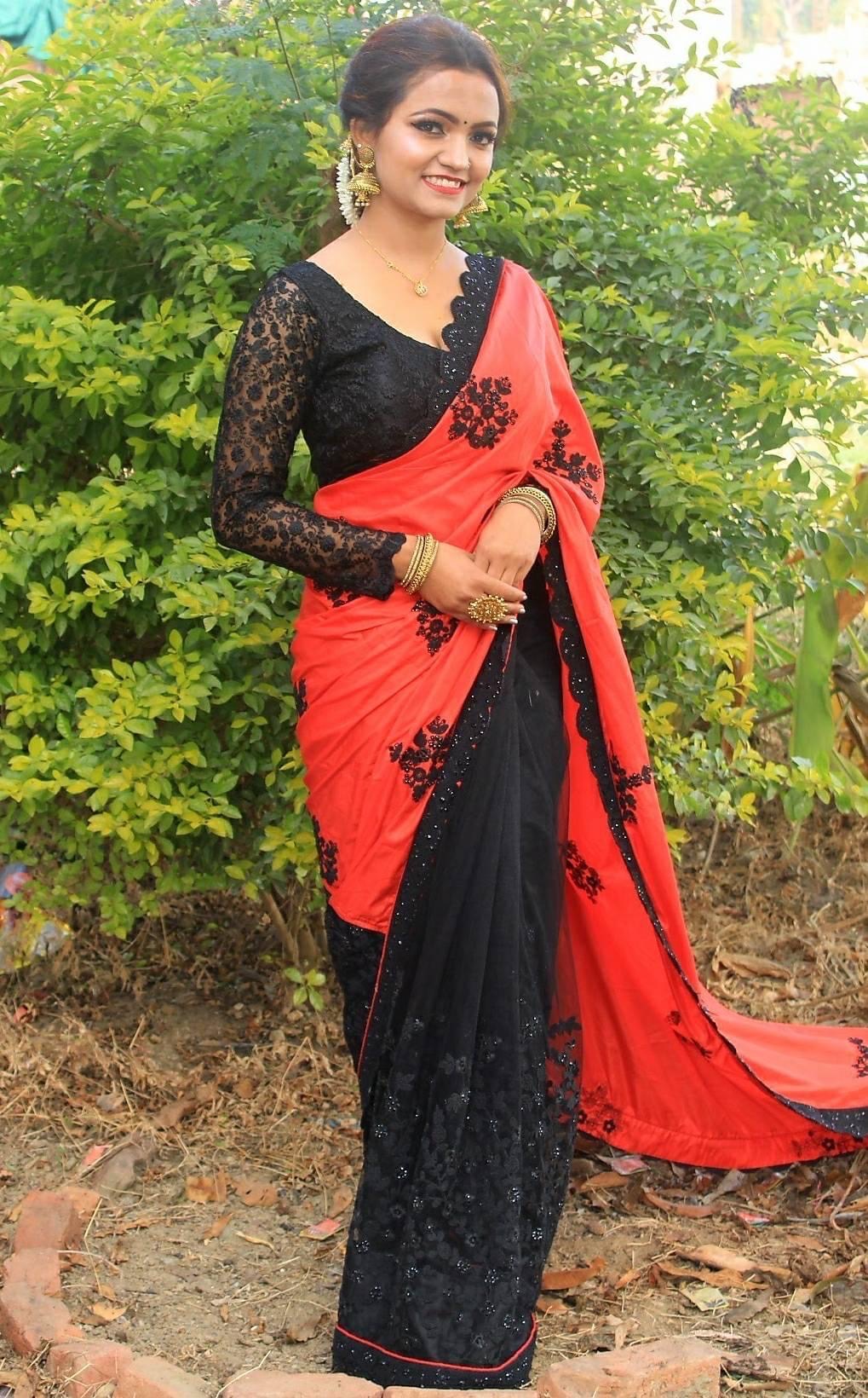 A woman stands outdoors in front of green bushes, wearing a black and red saree with black floral patterns, gold bangles, and large earrings. She is smiling and has her hands clasped in front of her.