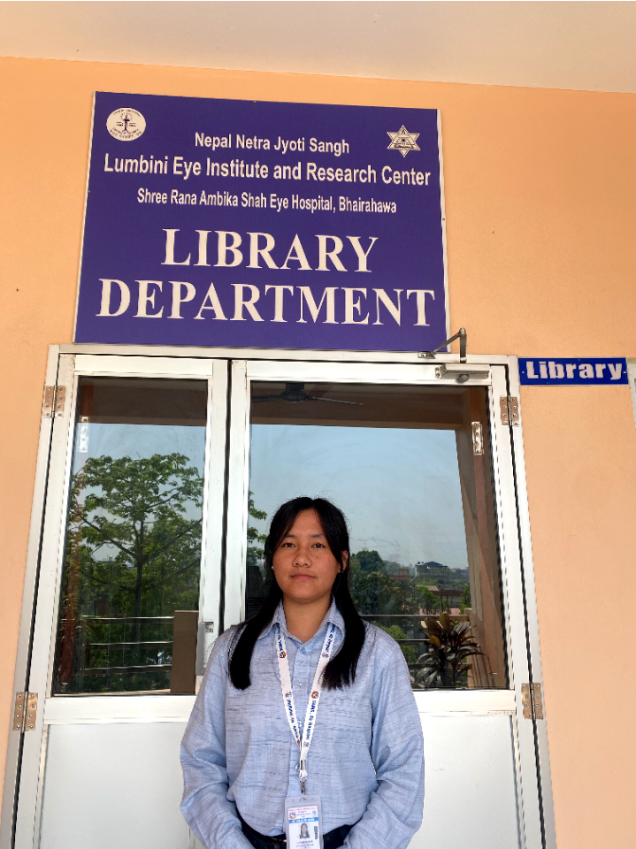A young woman stands in front of the Library Department entrance at Lumbini Eye Institute and Research Center, Bhairahawa. She wears a blue shirt and ID badge, with a blue and white sign above the door.