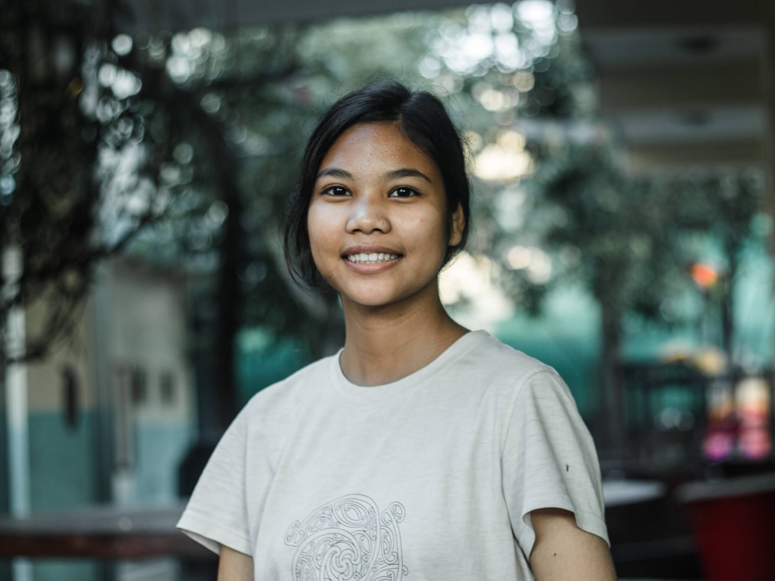 A young woman with dark hair tied back, wearing a light-colored t-shirt, smiles at the camera while standing outdoors with greenery and blurred buildings in the background.