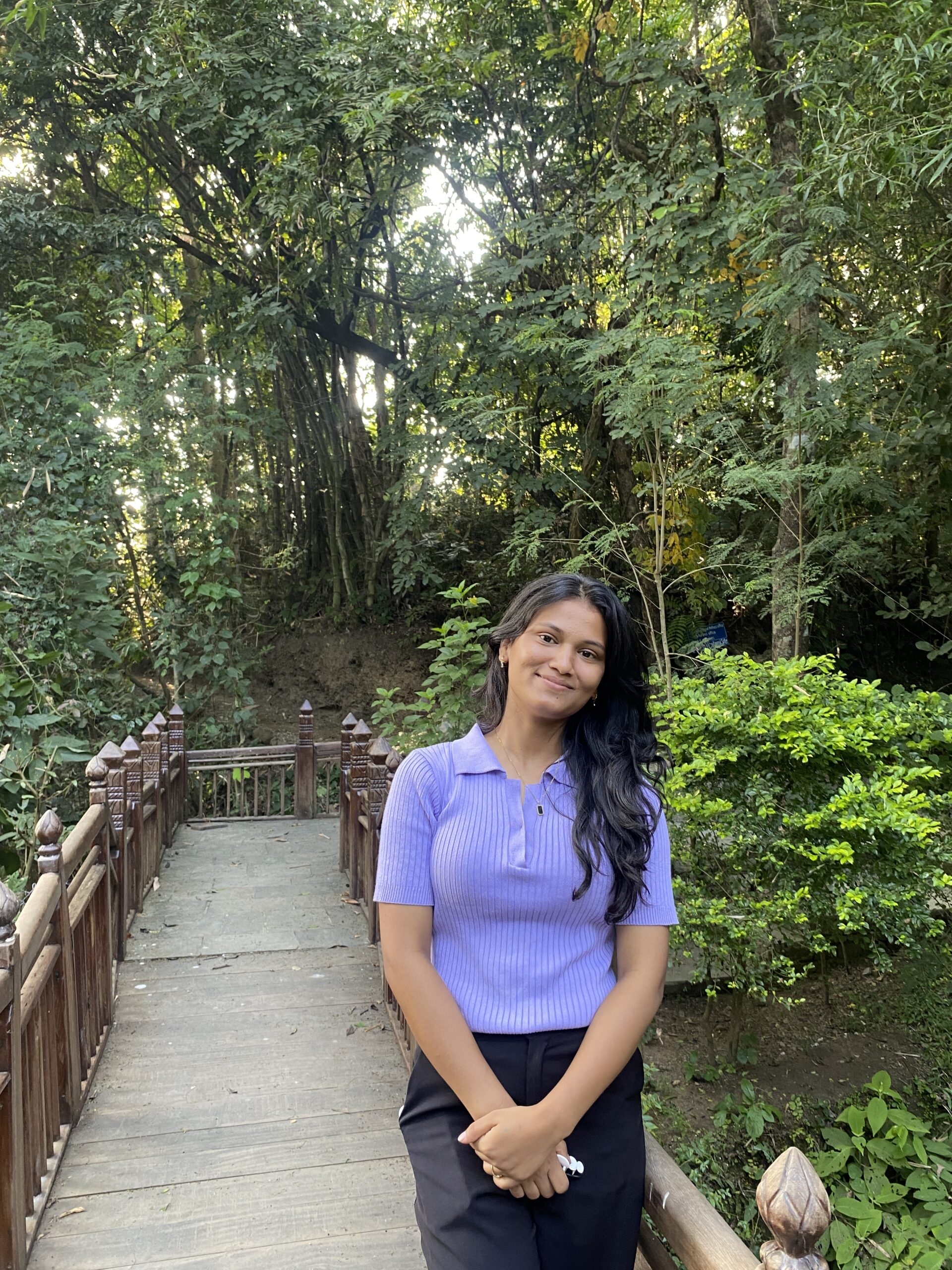 A woman with long dark hair, wearing a light purple shirt and black pants, stands smiling on a wooden bridge surrounded by lush green trees and foliage.