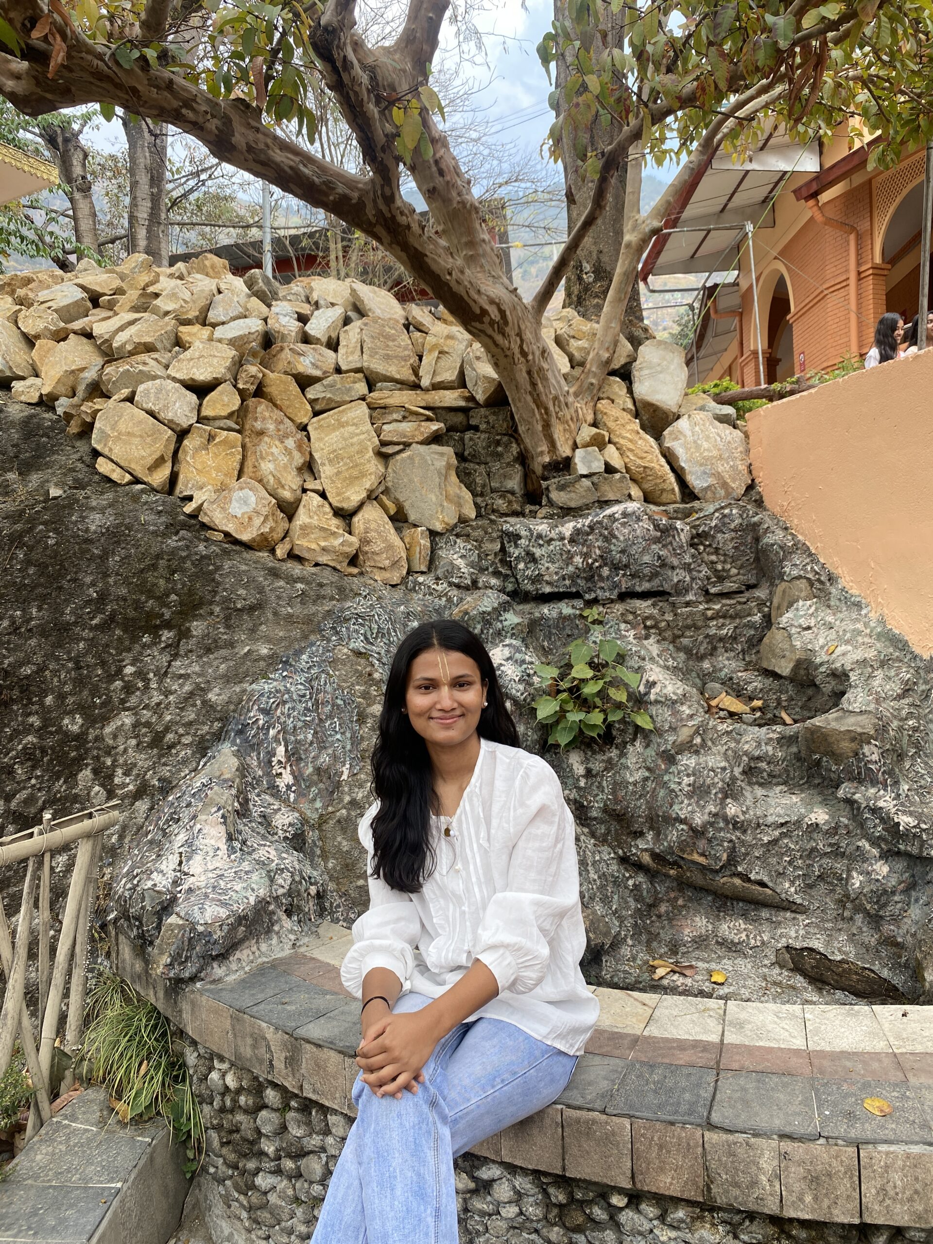 A woman with long dark hair, wearing a white top and light blue jeans, sits on a stone bench in front of a rocky slope with stacked stones and trees in an outdoor setting.