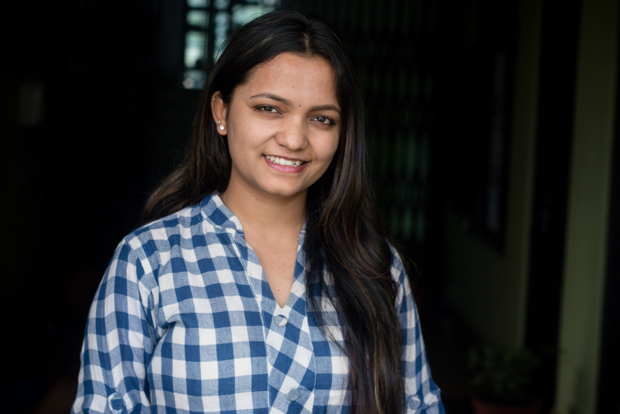 A young woman with long dark hair, wearing a blue and white checkered shirt, stands indoors and smiles at the camera. The background is softly lit and out of focus.