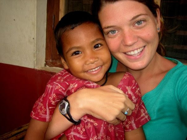 A smiling woman in a green shirt hugs a young child in a red patterned dress. They are both looking at the camera and appear happy, sitting together in front of a window and wall indoors.