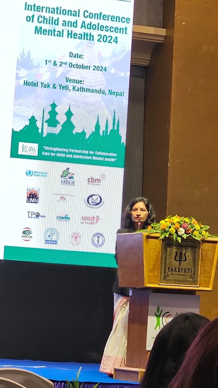 A woman speaks at a podium decorated with flowers during the International Conference of Child and Adolescent Mental Health 2024, held at Hotel Yak & Yeti in Kathmandu, Nepal. A large event banner is displayed behind her.