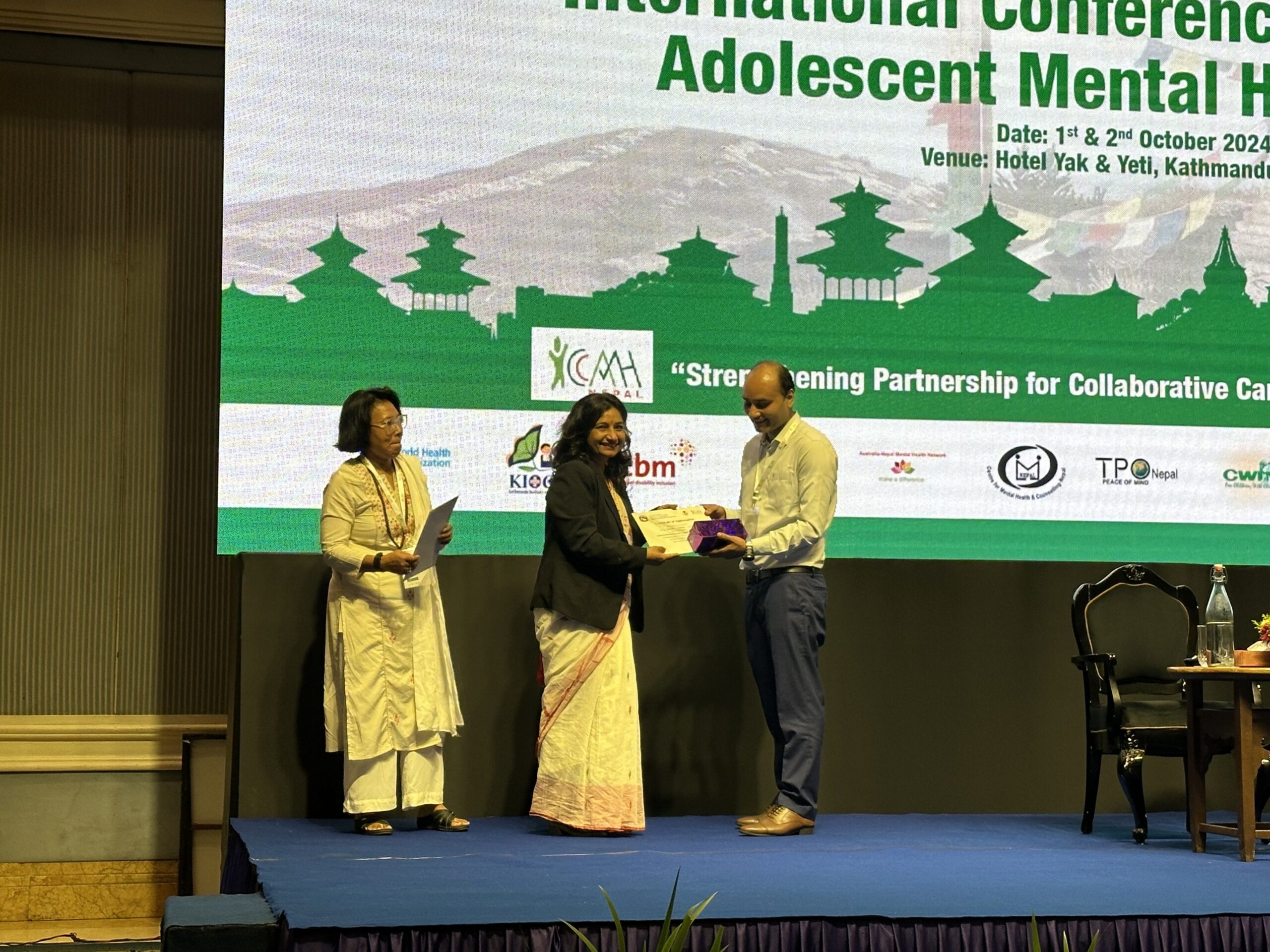 Three people stand on a stage at a conference. One woman is receiving an award from a man, while another woman observes. A presentation slide behind them displays event details about adolescent mental health.