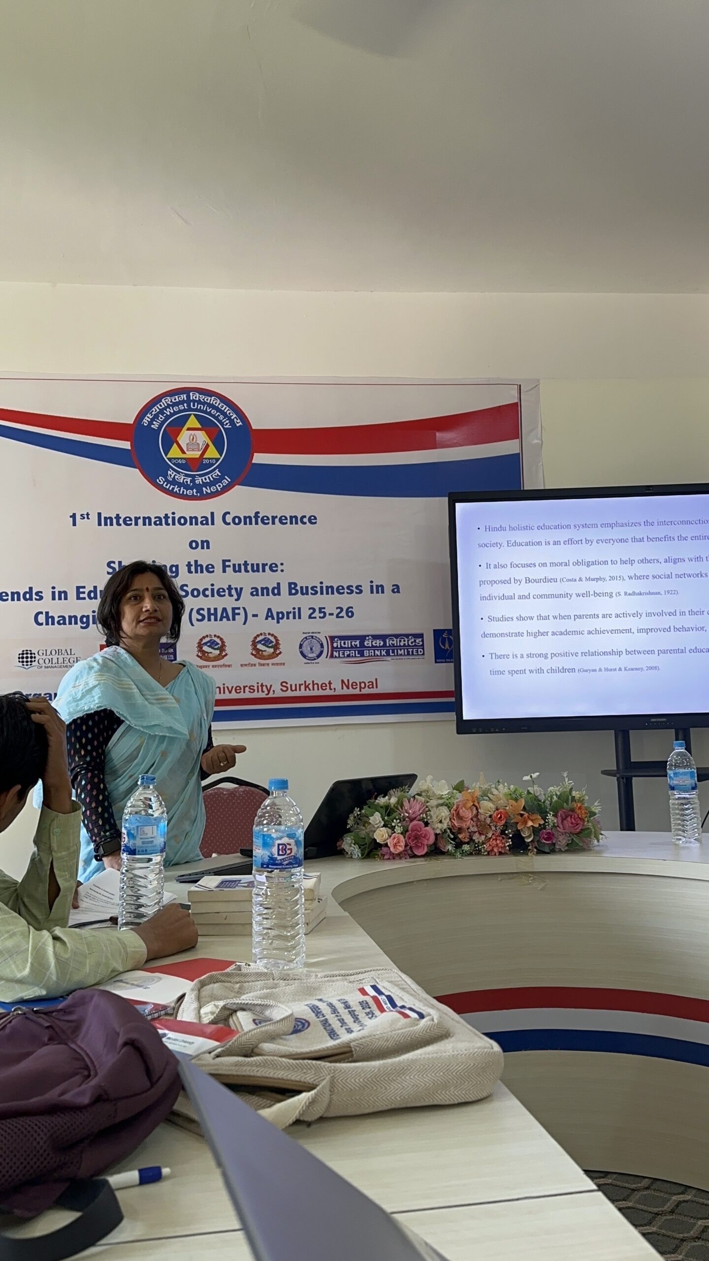 A woman in a blue sari presents at a conference, standing near a screen displaying text. Bottled water, bags, and flowers are on the table. A conference banner is visible behind her.
