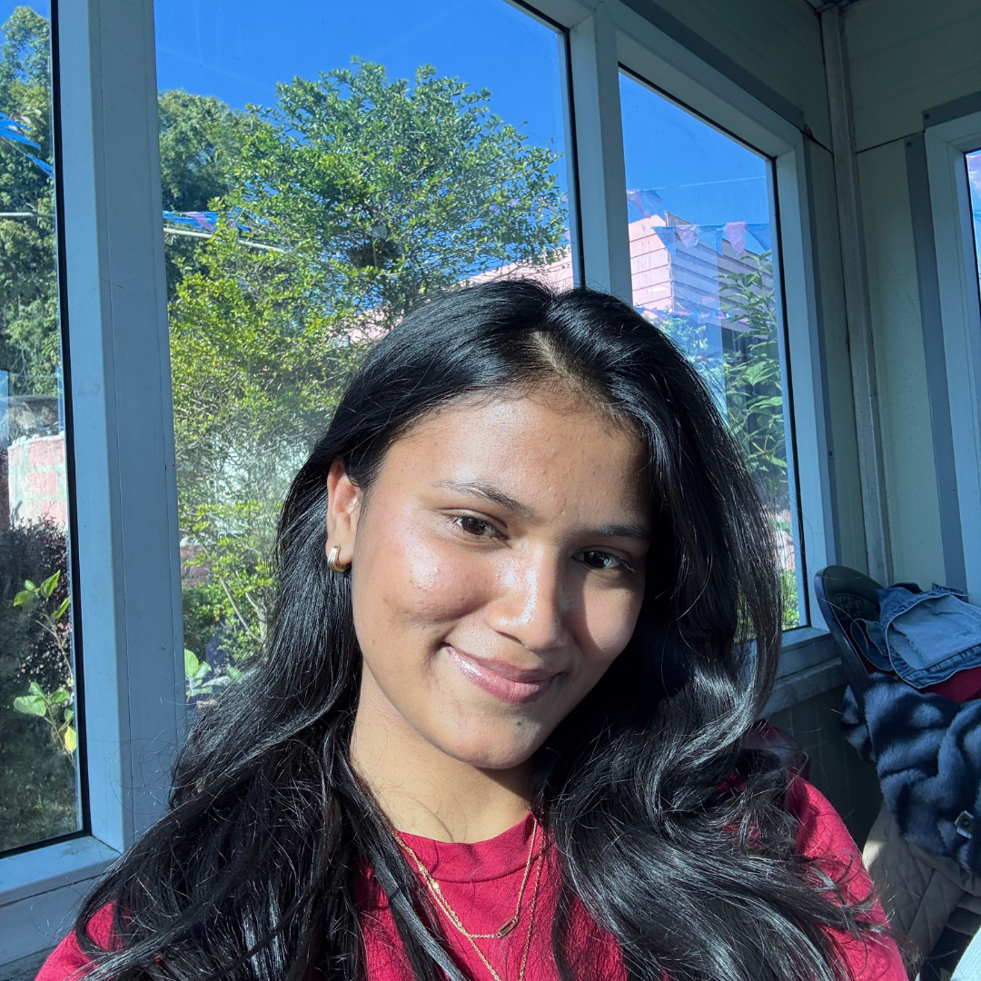 A young woman with long dark hair and a red shirt smiles at the camera, sitting indoors near large windows. Sunlight streams in, illuminating her face. Trees and a blue sky are visible outside.