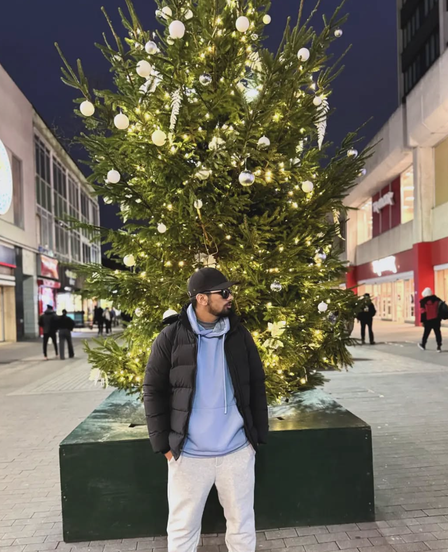 A man in casual winter clothing stands in front of a large, decorated Christmas tree with white lights and ornaments in an outdoor shopping area at dusk.