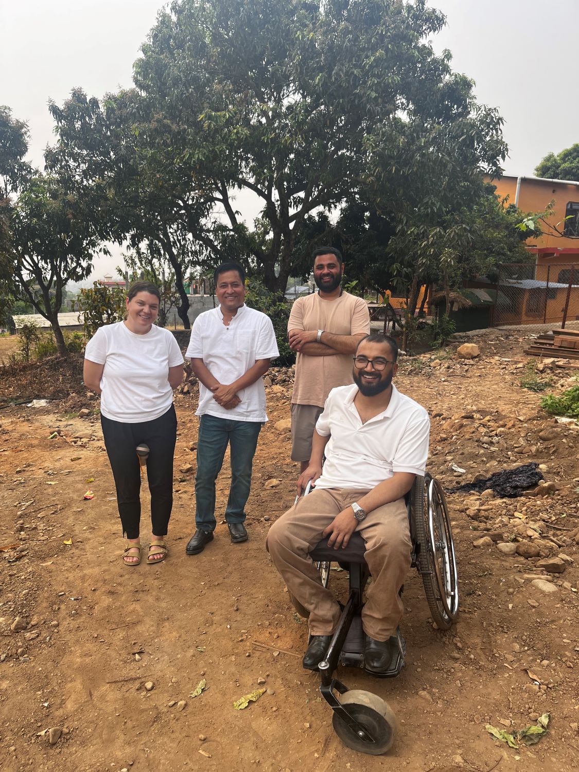 Four people are outdoors on a dirt area with trees and a building in the background; three are standing, and one person is sitting in a wheelchair. All are smiling and dressed in casual, light-colored clothing.
