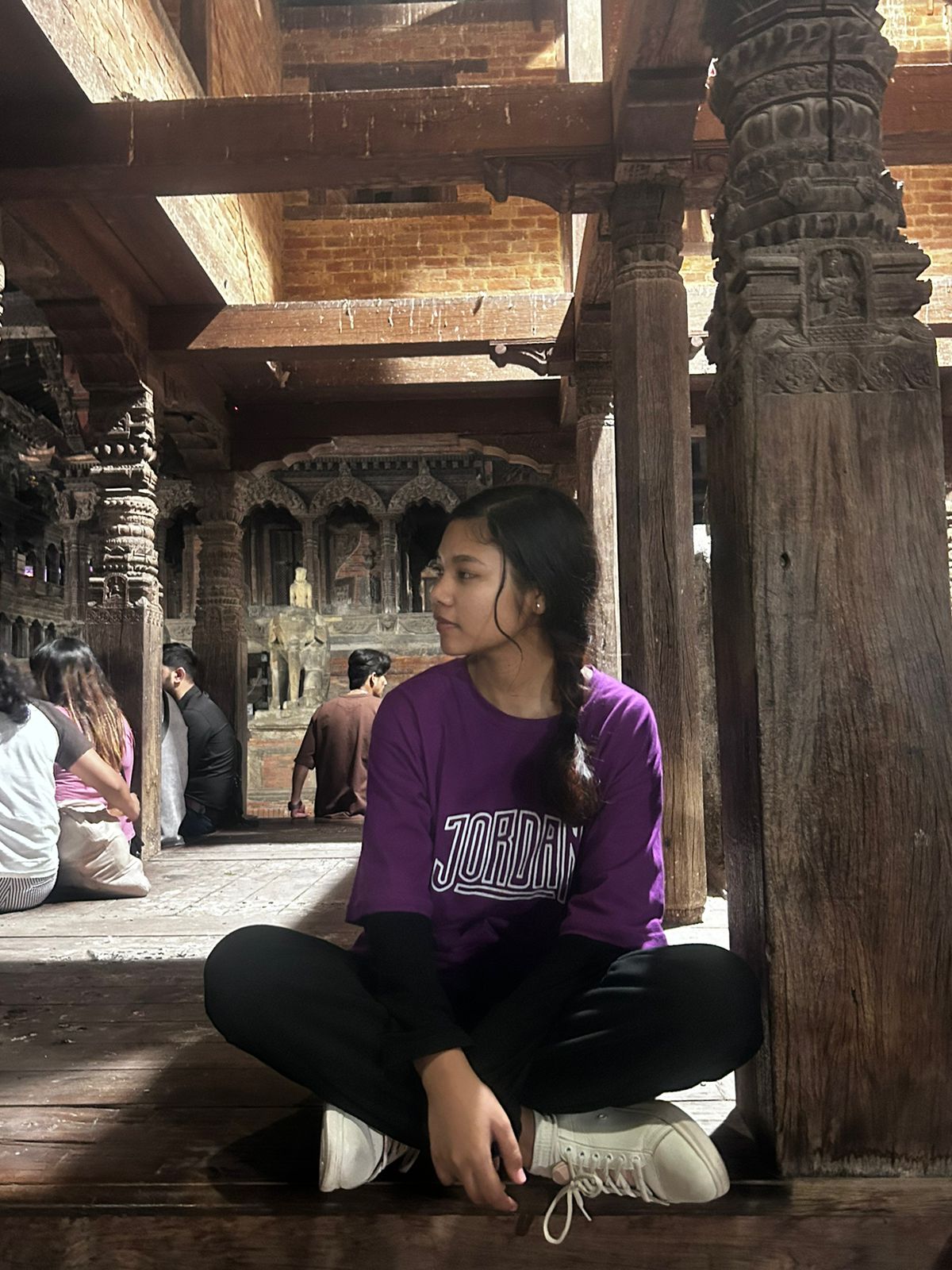 A young person wearing a purple "Jordan" shirt sits cross-legged on a wooden floor, looking to the side. Ornate wooden pillars and carvings surround them inside a historic building. Other people sit in the background.