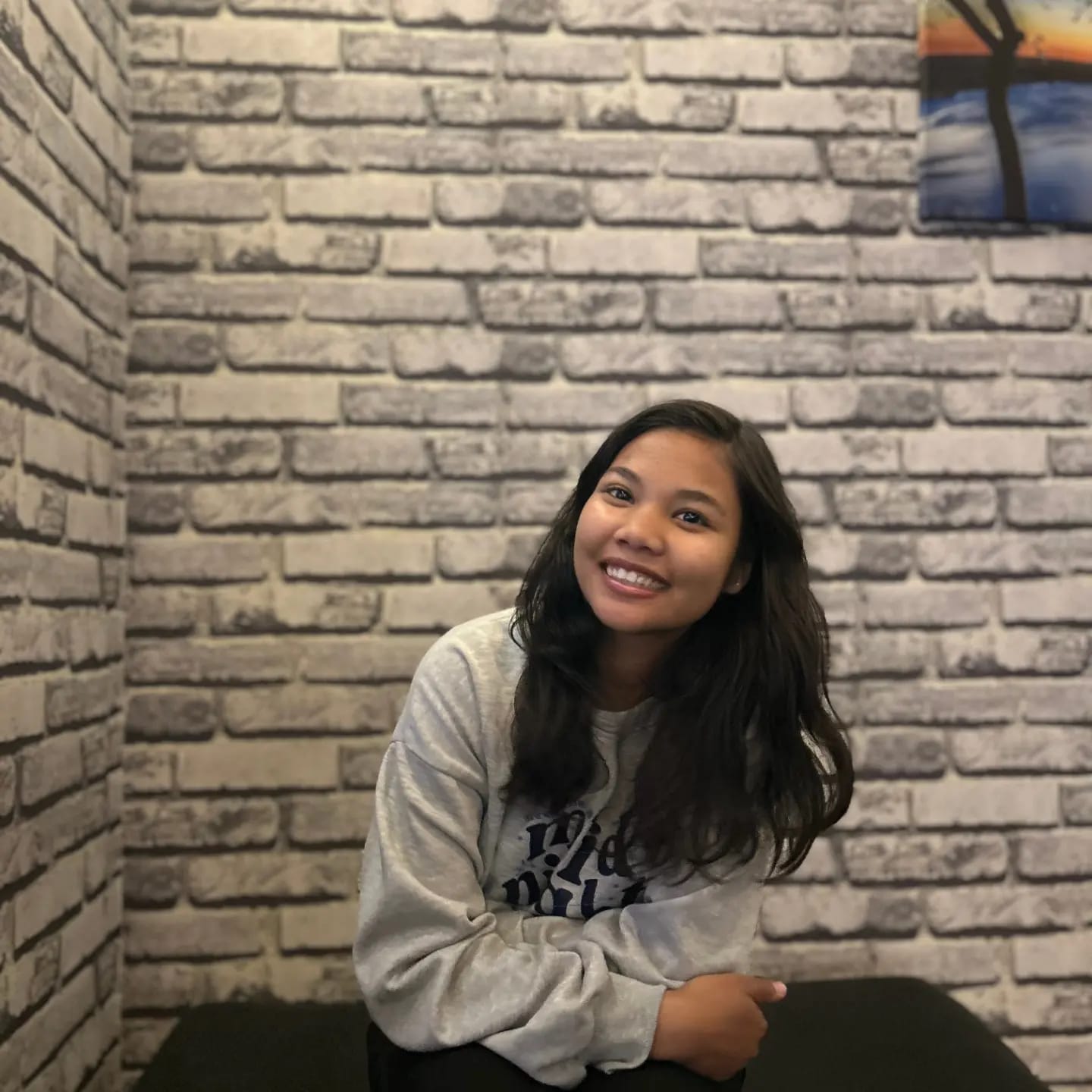 A young woman with long dark hair, wearing a gray sweatshirt, sits and smiles in front of a white brick wall with a painting in the upper right corner.