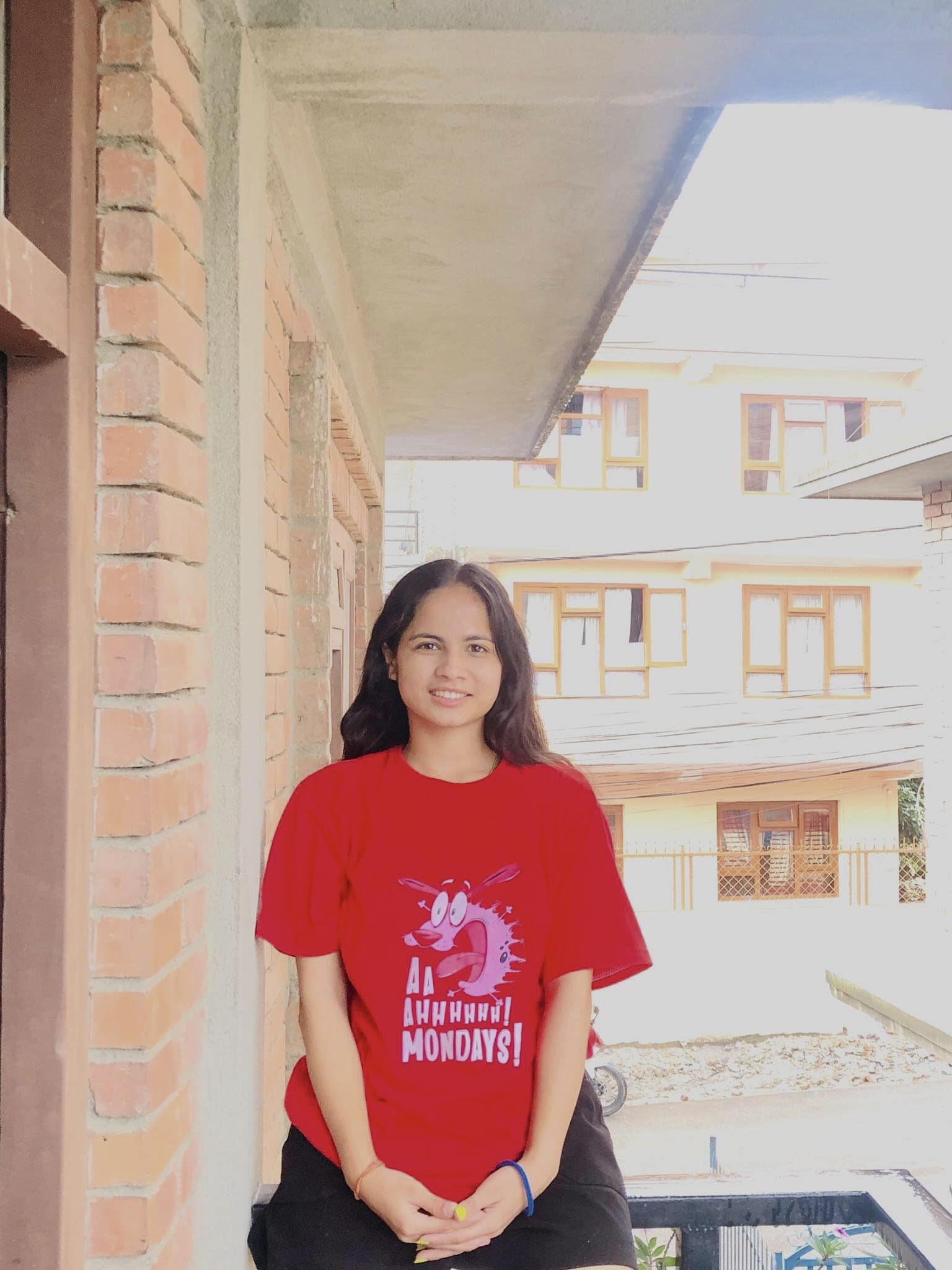 A young woman with long dark hair, wearing a bright red t-shirt with a cartoon and the words "Ahhhhhh! Mondays!", smiles while sitting in a corridor with brick walls and buildings visible in the background.