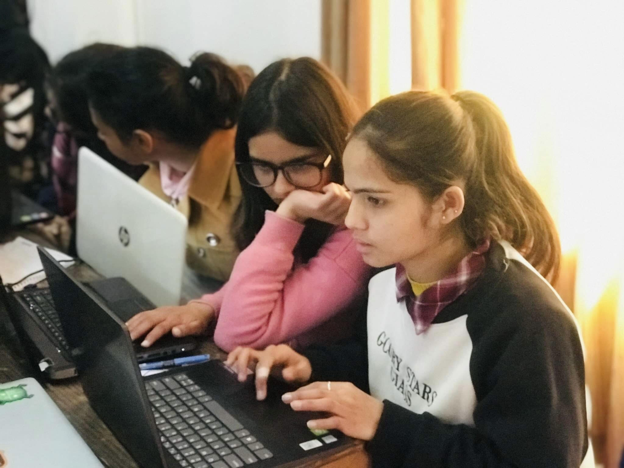 Three young women sit side by side at a table, working intently on laptops. Two focus on one screen, while another works independently. The setting appears to be a classroom or study area with natural light.