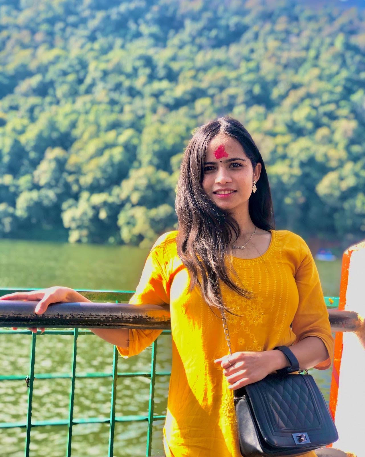 A woman with long dark hair, wearing a yellow kurta and black purse, stands by a railing in front of a green lake with forested hills in the background. She has a red tilak on her forehead and is smiling at the camera.
