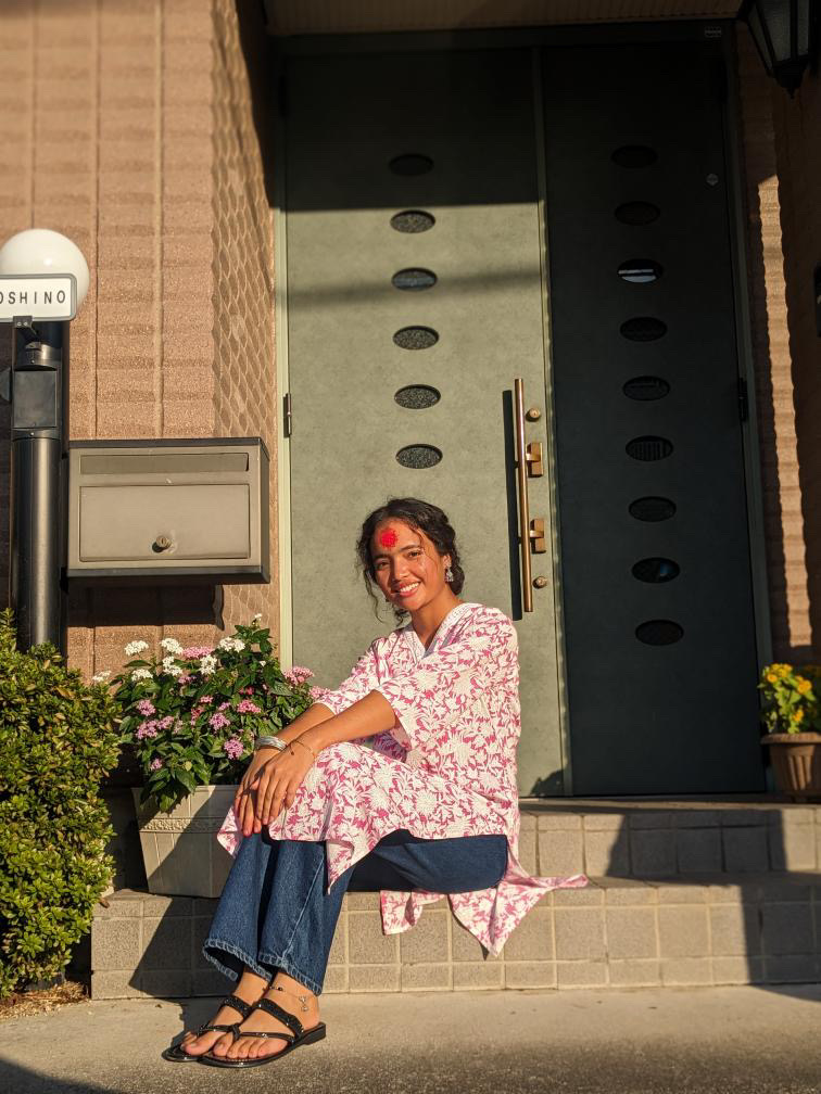 A smiling woman wearing a floral kurta, jeans, and sandals sits on steps outside a door, with sunlight on her face and flowers beside her. She has a red mark on her forehead and hair tied back.