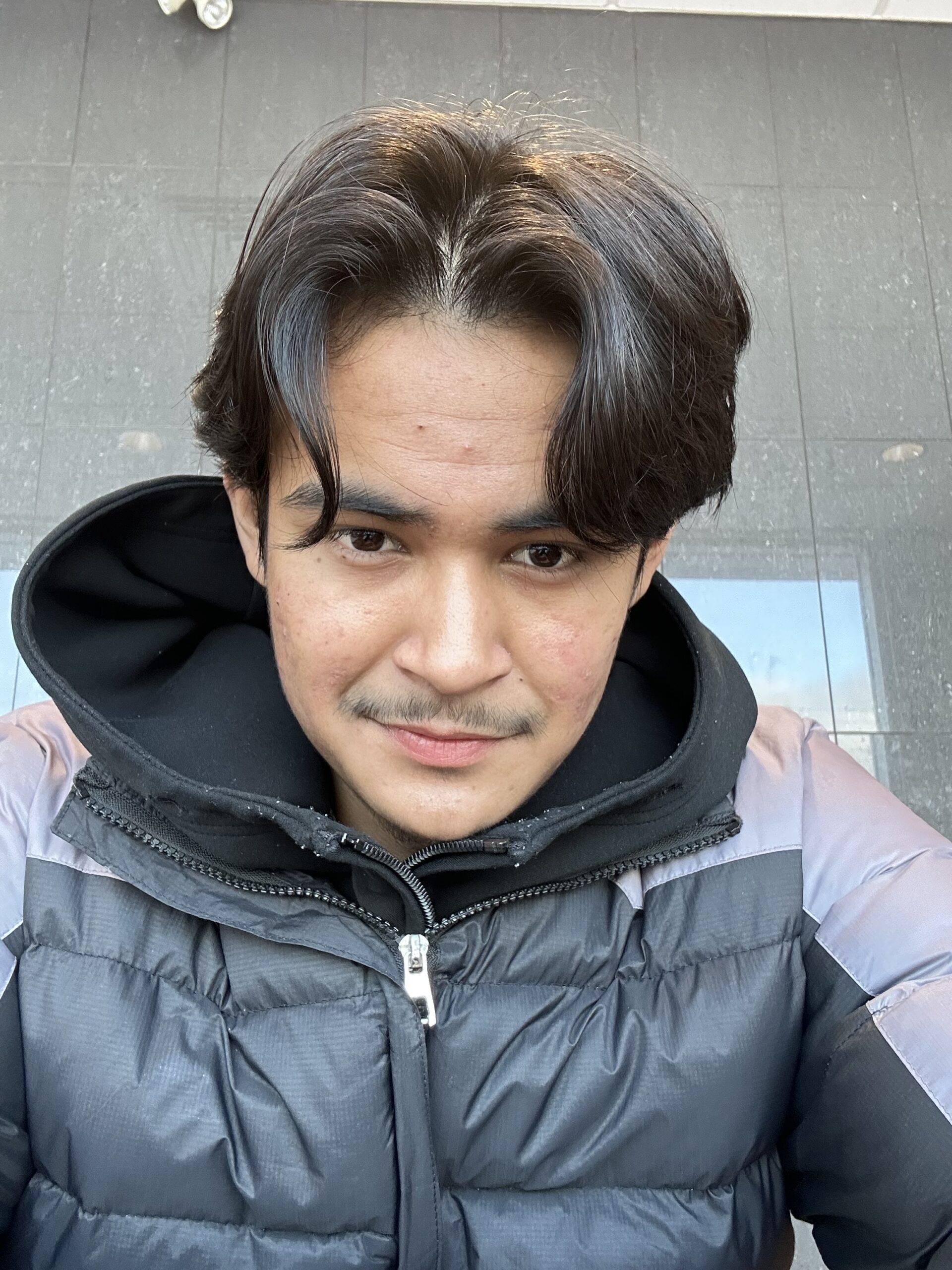 A young man with dark hair and a mustache wears a black and gray puffer jacket and a black hoodie, looking at the camera with a slight smile, standing against a reflective tiled wall.