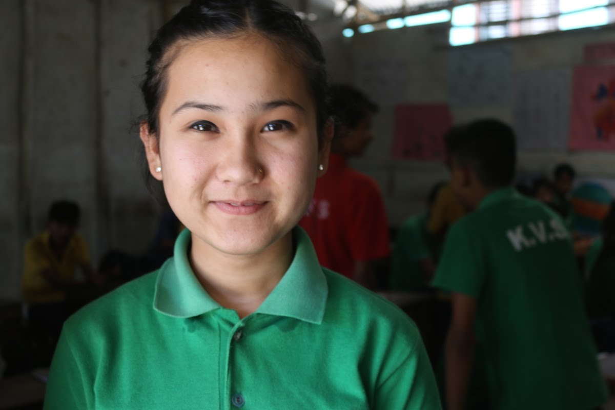 A young girl wearing a green polo shirt is smiling inside a classroom, with other students and posters visible in the blurred background.