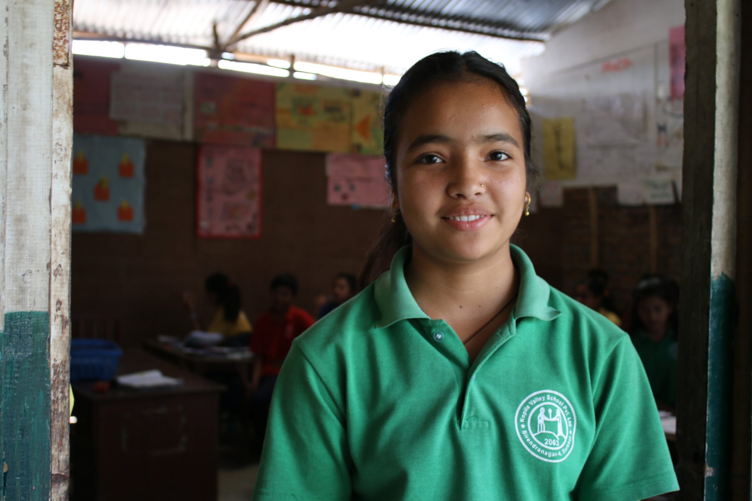 A girl wearing a green school uniform smiles while standing in a classroom doorway. Behind her, students sit at desks and colorful posters hang on the classroom walls.